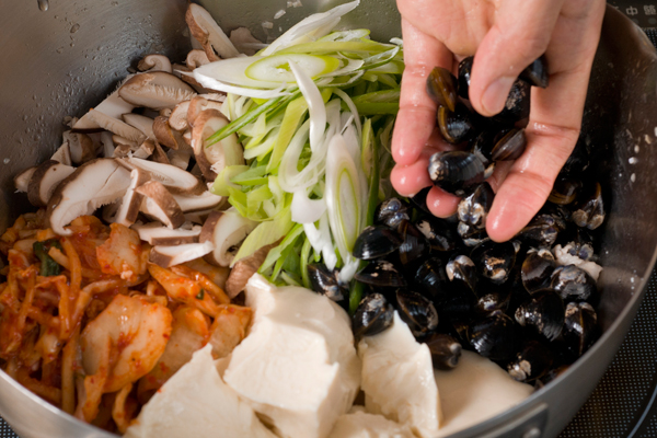 鍋にゴマ油とニンニクのみじん切りをいれ、豚バラ肉を炒める。さらに、豆腐をスプーンですくいながら入れ、白菜キムチ、1のシイタケ、ネギの半量、シジミを入れ、Ａを加えて煮込む。